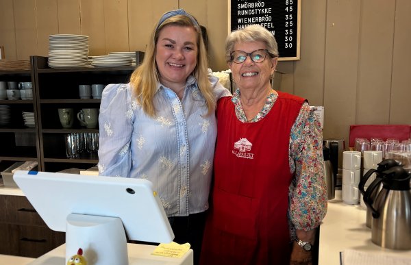 To kvinner står i en kafe bak et kassaapparat og smiler. Kvinne til venstre har blå bluse og blond hår, kvinne til høyre har briller og rød kjole.