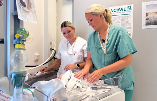 Kvinne med langt blondt hår samlet i strikk bak, iført turkis/grønn arbeidsuniform, står og undersøker en nyfødt baby. Ved siden av observerer en kollega med lyst hår samlet i strikk, iført hvit arbeidsuniform.