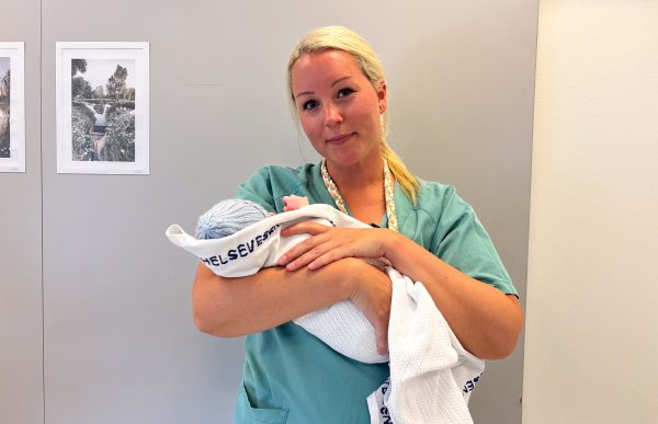 Kvinne med langt blondt hår samlet i strikk bak, iført turkis/grønn arbeidsuniform, holder en nyfødt baby pakket i hvit pledd foran kamera.
