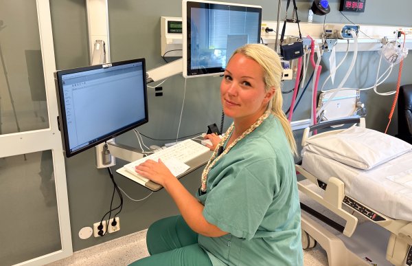 Kvinne med langt hår samlet i strikk bak, iført turkis/grønn arbeidsuniform, sitter foran en skjerm på pasientrommet for å dokumentere arbeidet. I bakgrunnen ses et sykehusrom.