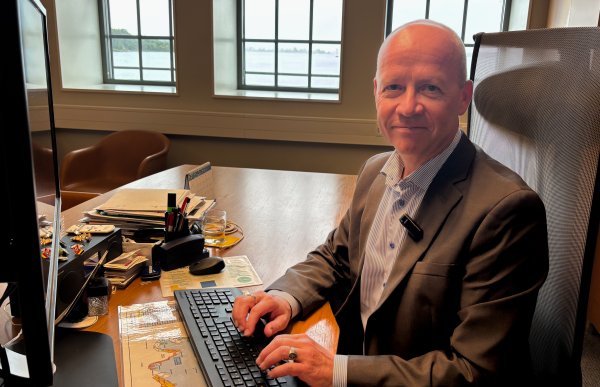 Mann i mørk grå dressjakke og blå skjorte sitter ved datamaskin på kontor. Han smiler til kamera, med vinduer mot havet i bakgrunnen.