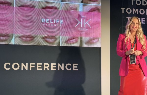 Kvinne med langt blondt hår, iført rosa dressjakke og langt rødt skjørt, står foran en scene og holder et foredrag.