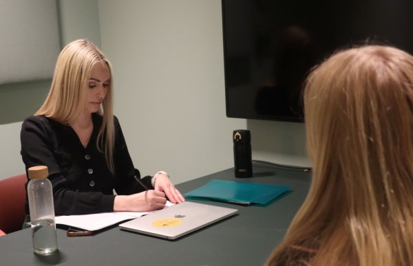 En kvinne med langt lyst hår og svart overdel skriver noe på en blokk. En annen kvinne sitter med ansiktet mot kamera. Hun har også langt lyst hår. Mellom kvinnene er det et bord.