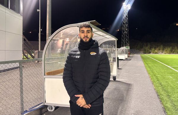 Mann med svart hår, skjegg og bart poserer foran en fotballbås utendørs om kvelden. Han har på seg en langermet jakke, og lys fra stolper skimtes i bakgrunnen.