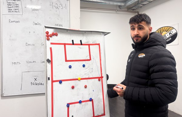 Mann med svart hår, skjegg og bart står i garderobe og holder en tusj foran markerte tavler. Han har på seg en langermet jakke