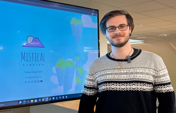 Mann med kort, mørkt hår og briller står foran en TV-skjerm på et kontor. Han har på seg en langermet genser i svart og grått. I bakgrunnen skimtes et felles kontorområde.