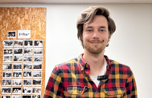 Mann i rutete rød- og gulskjorte poserer foran kamera. I bakgrunnen skimtes en vegg med bilder fra kontorlandskapet.