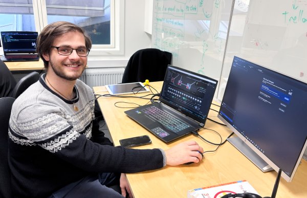 Mann med kort, mørkt hår, briller og langermet genser i svart og grått sitter foran en PC-skjerm på kontoret. Han vrir hodet og smiler til kamera. I bakgrunnen skimtes et kontorlandskap.
