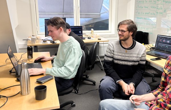 Mann med kort, mørkt hår, briller og langermet genser i svart og grått sitter sammen med teamet sitt og ser på skjermen til en kollega. Kollegaen, iført turkis genser, er fokusert på programmering, mens en annen kollega i rutete rød- og gul skjorte ser på og smiler til kamera.