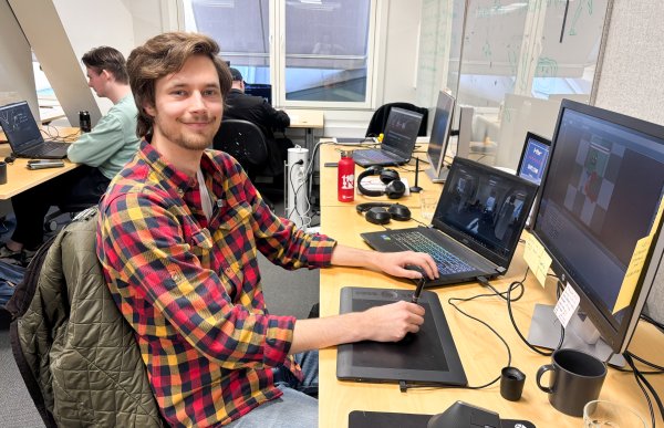 Mann med brunt hår, iført rød-gul rutet skjorte, sitter foran PC-en og animerer. Han smiler mot kamera, med den ene hånden på animasjonsbrettet og den andre på PC-en. I bakgrunnen skimtes kontorlandskapet og et glimt av en kollega.
