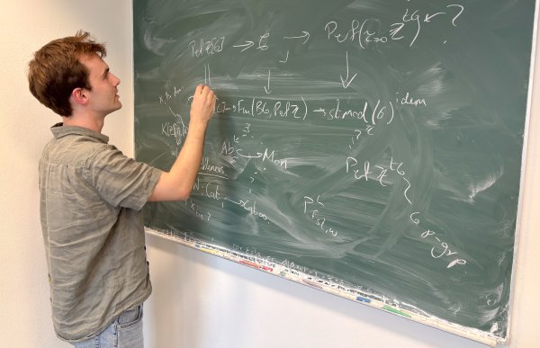 Mann med brunt, kort hår og grønn, kortermet t-skjorte står foran en grønn tavle og skriver matematiske formler.