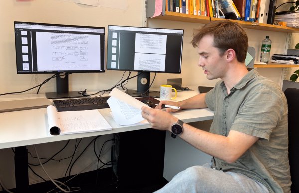 Mann med brunt, kort hår og grønn, kortermet t-skjorte sitter og leser forskning. I bakgrunnen vises kontoret hans med dataskjermer.