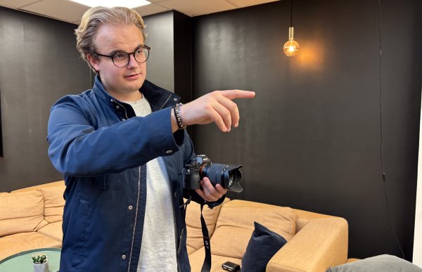 Videokunstner med kamera under opptak Mann med blondt hår og briller, iført grå T-skjorte og marineblå jakke, holder et kamera og gir instruksjoner under et oppdrag. I bakgrunnen sees et sofamiljø.