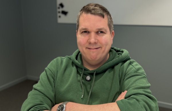 En mann sitter ved et bord med armene krysset foran seg og ser mot kamera med et svakt smil. Han har kort, gråsprengt hår og er kledd i en grønn hettegenser. På håndleddet har han en klokke med metallrem. Bak ham er en grå vegg med et enkelt kunstverk i hvitt og svart.