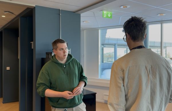 To menn står og snakker sammen i et moderne kontorlokale med store vinduer og utsikt. Mannen til venstre, kledd i grønn hettegenser, vender seg mot den andre og ser konsentrert ut. Den andre mannen står med ryggen til og lytter. Han har briller og en lys skjorte på seg. Over dem henger et grønt nødutgangsskilt i taket.