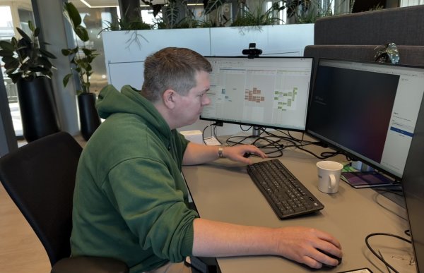 En mann sitter ved en kontorpult og arbeider ved to dataskjermer. Han holder hånden på en datamus og ser konsentrert på skjermen foran seg. På bordet står en kaffekopp, og bak skjermene er det planter og kontorinnredning. Skjermene viser ulike programvinduer og oversikter.