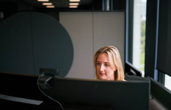 En kvinne med lyst hår sitter og ser på en PC-skjerm. Hun har ansiktet mot oss, men ser konsentrert ut. I bakgrunnen er det vinduer og flere kontorplasser.