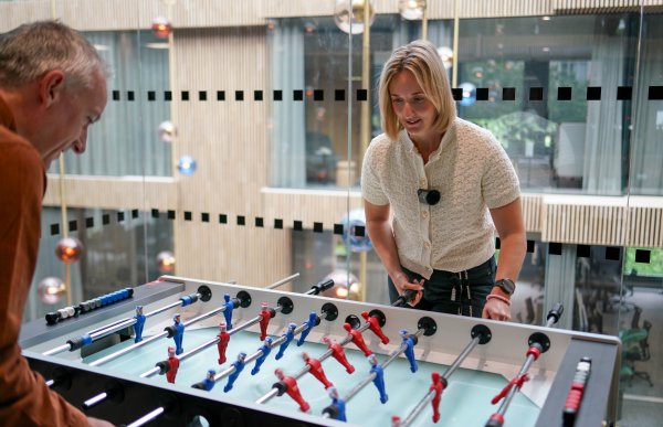 En blond kvinne og en mann med grått hår spiller bordfotball mot hverandre. Det er blå og røde menn på bordspillet. Begge personene ser ivrig og konsentrerte ut.