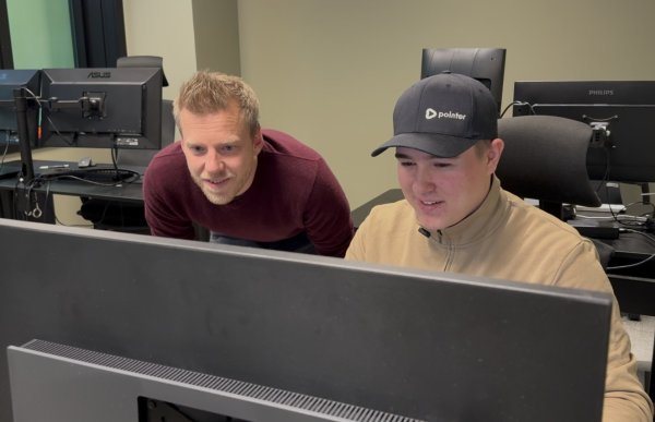To menn ser på en dataskjerm på et kontor. Den ene sitter foran skjermen og har på seg svart caps og beige genser. Den andre står bøyd ved siden av ham og har kort, lyst hår og en mørkerød genser. I bakgrunnen er flere dataskjermer.