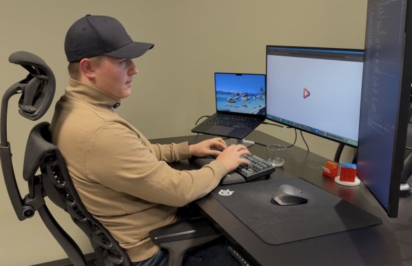 En ung mann med svart caps, beige genser og blå jeans sitter i en kontorstol og arbeider ved et skrivebord. Han skriver på et tastatur foran to store dataskjermer, hvorav den ene viser programmeringskode. Ved siden av skjermene står en bærbar PC.