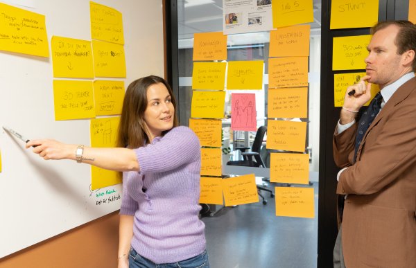 Kvinne med langt, brunt hår i lilla cardigan og mann i en brun dressjakke. Kvinnen peker med en tusj på en tavle med gule post-it-lapper, mens hun ser på mannen. Mannen i dress står ved siden av henne og lytter.