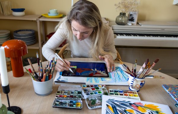 Ei dame med langt og lyst hår. Hun har hvit genser på seg. Hun tegner på en iPad. Foran henne ligger tegnesaker og et fargerikt bilde. Hun er i en leilighet.