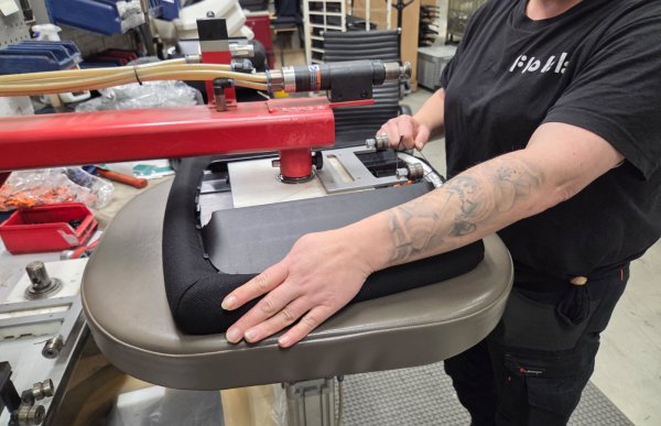 Vi ser nedre del av en kvinne i sorte klær. Hun holder baksiden av et sete i hendene og et apparat i rød farge er festet på setet. Setet hviler mot en slags krakk. Det ser ut som hun er i et slags verksted, og i bakgrunnen er det mye forskjellig utstyr.