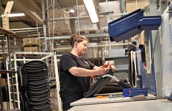 En kvinne med svart antrekk står ved en arbeidsbenk på en fabrikk og jobber med en stolseterygg. Personen bruker en håndholdt stiftemaskin for å feste trekk. På benken ligger verktøy og briller, og i bakgrunnen sees hyller og reoler med lagerkasser og stoler stablet i høyden. Arbeidsområdet er godt opplyst med en lampe over benken.