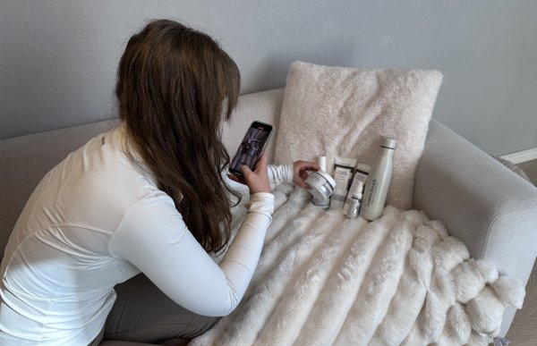Influenser Lisa Semb lager et innlegg om en hudpleieserie. En kvinne med langt brunt hår sitter i en lys sofa og tar et bilde med mobilen av flere hudpleieprodukter som ligger på et mykt, hvitt teppe og står lent mot en pute. Ved siden av produktene er en grå drikkeflaske. Rommet har lyse vegger.