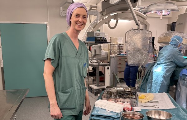 En kvinne i grønne operasjonsklær og lilla operasjonshette står i et operasjonsrom og smiler mot kamera. Foran henne står et bord med kirurgiske instrumenter, skåler og sterile duker. I bakgrunnen jobber annet helsepersonell ved en pasient som ligger på operasjonsbordet, omgitt av avansert medisinsk utstyr og operasjonslamper.