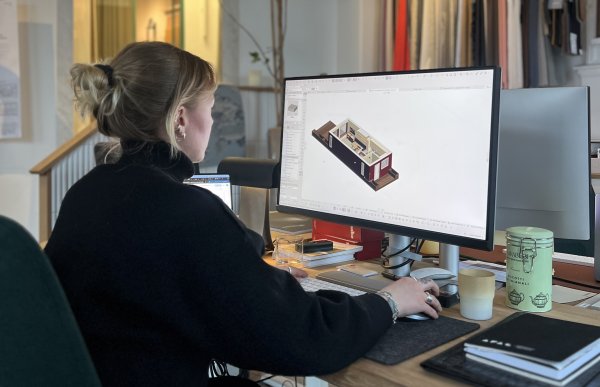 En dame sitter med ryggen til ved en kontorpult og jobber med noe på en pc-skjrm. På skjermen er et utsnitt av et hus