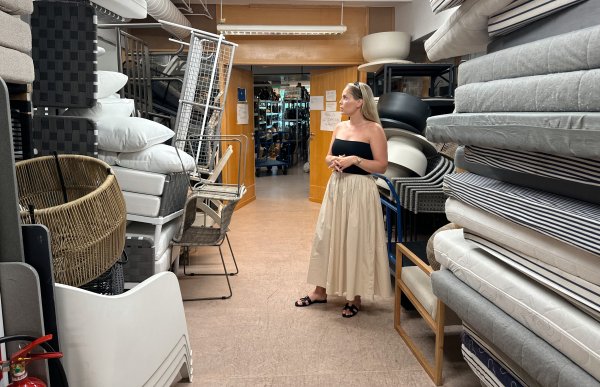 En dame med blondt hår, svarte solbriller på hodet, svart topp og beige skjørt står og titter på noen lyse, stabla puter til utemøbler.