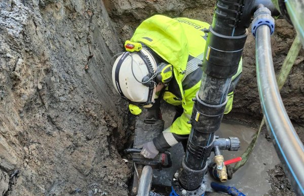 En anleggsrørlegger står dypt nede i en smal gravegrop omgitt av våt jord. Han jobber med et lekk rør der vann siver ut, mens store rør og koblinger ligger montert rundt området.