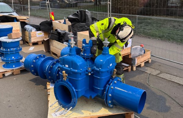 En anleggsrørlegger bøyer seg over en stor blå ventilblokk som ligger på en pall ute på en byggeplass. Rundt ham står trepaller, rørdeler og arbeidsutstyr. Bak ham står en boligblokk og et gjerde som sperrer av området.