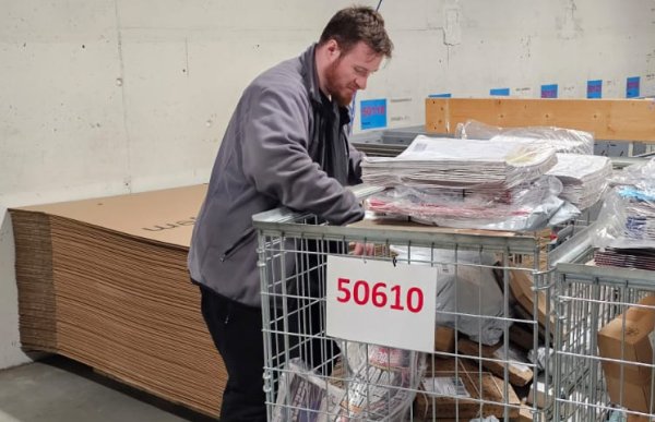 En mann i grå jakke står bøyd over en stor trådkurv med aviser, poser og pakker. Bak ham ligger flate papplater stablet langs veggen.