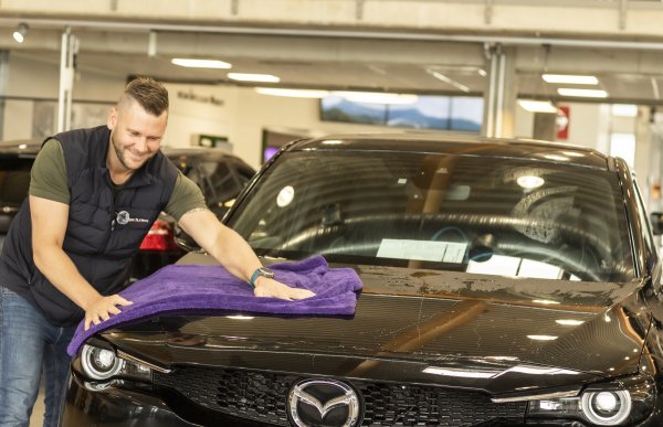 Mann i mørk vestog blå skjorte tørker over en sort mazda med et lilla håndkle inne hos en bilforhandler