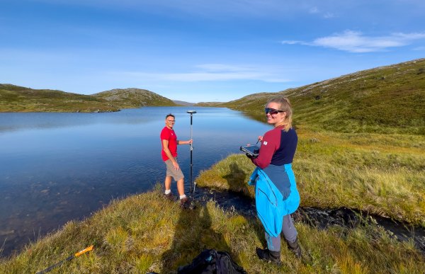 En kvinne med turantrekk i rødt og blått og en mann i rød t-skjorte og beige turshorts i står med måleutstyr ved et vann