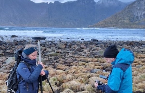 En kvinne med blå turjakke og blått pannebånd og en mann med blå lue og blå turjakkestår med måleutstyr ved sjøen 