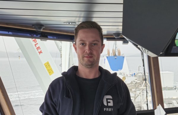 En mann i mørke klær står i et styrhus i en båt. Det er vinduer bak ham.