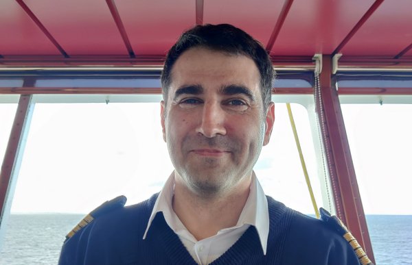 Portrett av kaptein Lars-Morten Grønmo. Han er avbildet på broa på båten, vi ser horisonten ut vinduene. Han er iført mørkeblå genser med tre gullstriper på hver skulder.