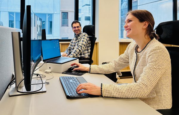 Produktleder Kari Clifford på kontoret.  En kvinne og en mann sitter foran hver sin dataskjerm i et lyst kontorlandskap. Begge smiler, og kvinnen ser opp fra tastaturet mens mannen ser mot henne. Store vinduer slipper inn dagslys bak dem.