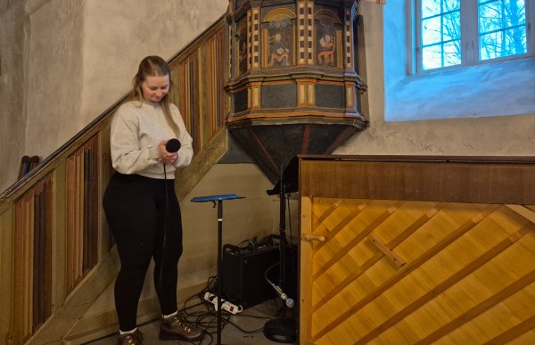 En ung kirketjener står ved siden av prekestolen i en gammel kirke. Hun holder en håndholdt mikrofon og ser ned på den mens hun gjør klart lydutstyr før et arrangement. Bak henne sees den utskårne, fargerike prekestolen, en bratt trapp med trepanel og et stort vindu som slipper kaldt blått dagslys inn i kirkerommet.