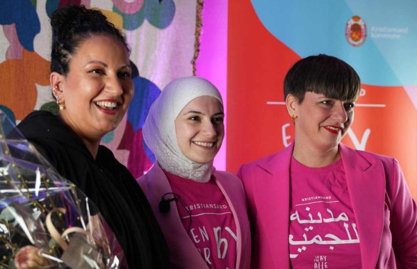 Tre kvinner står side om side og smiler. Den ene holder en blomsterbukett, den andre har på hijab, og den tredje er kledd i rosa jakke og T-skjorte. Bak dem henger et fargerikt teppe og en plakat fra Kristiansand kommune.