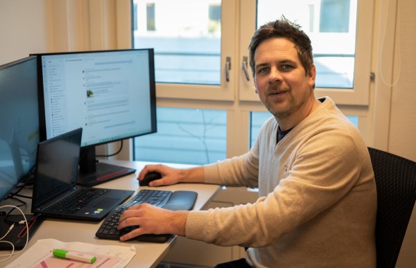 Mannen sitter ved en kontorpult foran to store dataskjermer og ser mot kamera. Han holder hånden på tastaturet, og vinduer bak ham slipper inn dagslys. Det ligger papirer og en markør på skrivebordet.