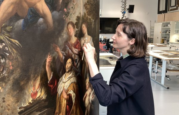 En dame med mørkt skulderkort hår står ved et maleri og bruker en liten børste på overflaten. I bakgrunnen vises et rom med forskjellig typer kunstutstyr. 