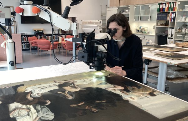 En dame sitter ved et bord med en forstørrelsesglass-maskin og titter på et maleri som ligger på bordet. I bakgrunnen vises en stolgruppe og andre verktøy innen kunstfag i et rom med skap  og kontorrekvisitta.  