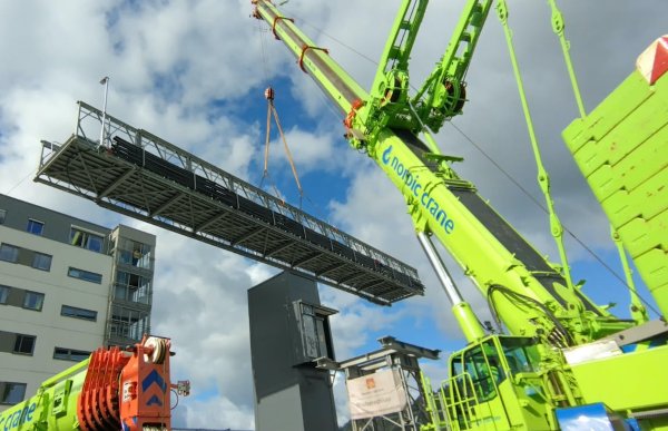 Kranfører Dorthe Sandvik gjør et løft med en bru. En grønn kran løfter en bru opp i lufta. I bakgrunnen ser man en hvit og grå bygning med vinduer. Det er flere grønne elementer i metall rundt krana. Brua er av metall med rekkverk på.