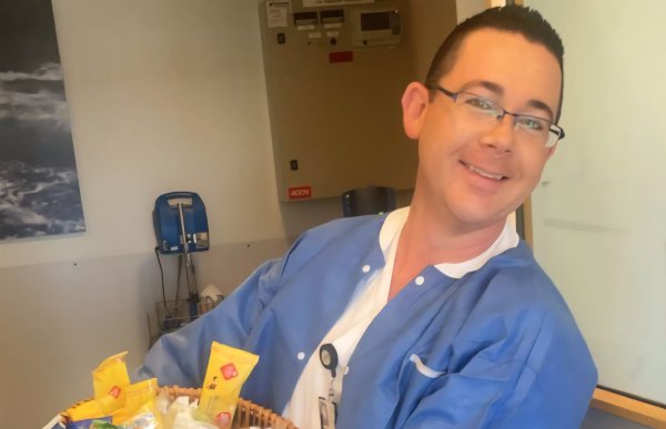 En mann i blå sykepleieruniform smiler mot kamera og holder en stor flettet kurv full av innpakkede mat- og snackprodukter. Han står inne på et rom med hvite vegger og et lite vindu til høyre.