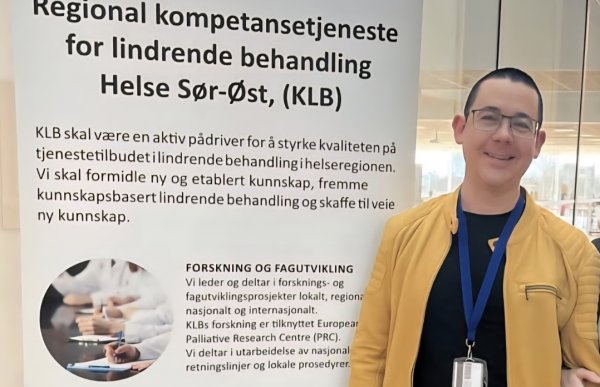 En mann med kort mørkt hår, briller og gul jakke står ved en informasjonsplakat.
