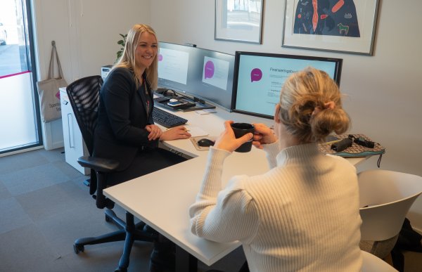 To kvinner sitter overfor hverandre ved et skrivebord i et kontor. Den ene sitter bak en pult med to skjermer, mens den andre sitter på en stol og holder en kopp. På veggen henger kunst, og kontoret fremstår ryddig og lyst.